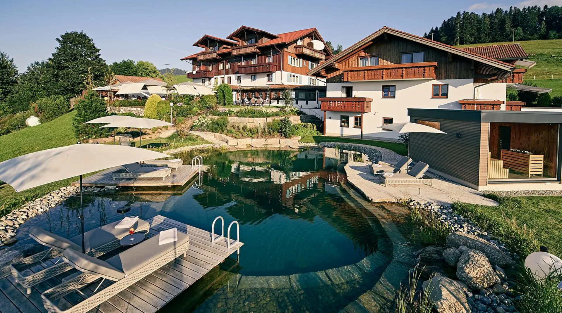 Hausansicht des Hotels Birkenhof in Oberstaufen mit Naturschwimmteich, Holzstegen, Liegen und Sonnenschirmen im Vordergrund.