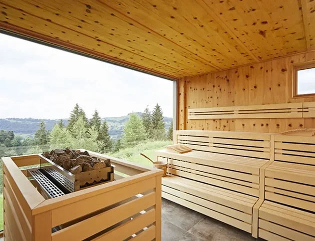 Panoramasauna im Hotel Birkenhof in Oberstaufen mit Blick ins Allgäu durch ein großes Fenster, helle Holzbänke und Saunaofen im Vordergrund.
