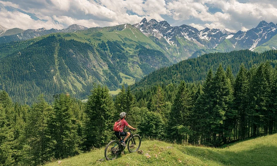 Mountainbiker mit rotem Rucksack fährt über eine Wiese inmitten dichter Wälder, mit Blick auf grüne Täler und schneebedeckte Gipfel der Allgäuer Alpen unter leicht bewölktem Himmel.