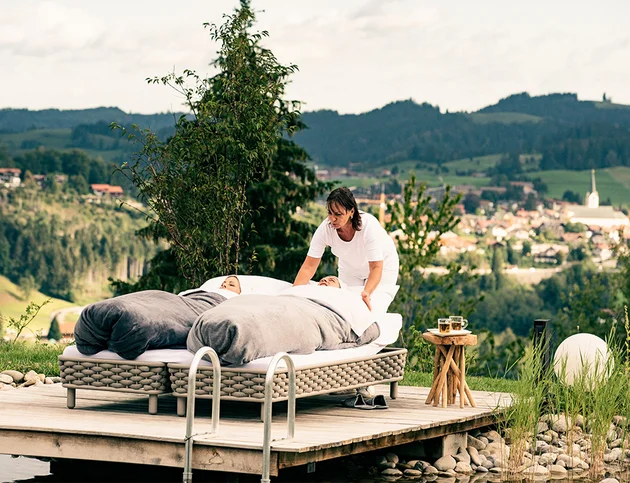 Zwei Personen liegen in Decken gehüllt auf Liegen am Steg, eine Frau in Weiß betreut sie, im Hintergrund Blick auf das Allgäu und Oberstaufen.