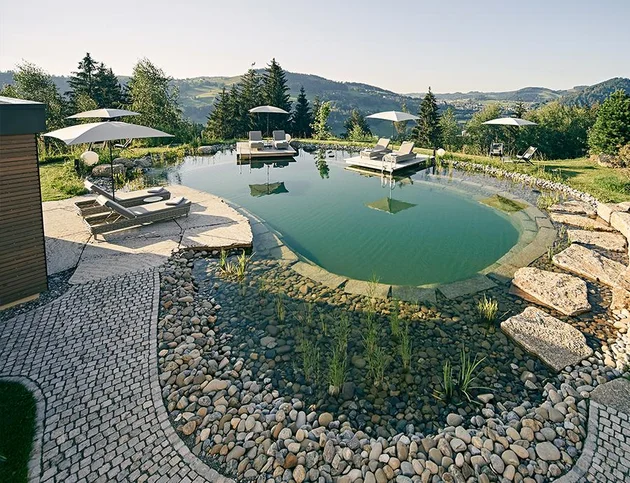 Naturschwimmteich des NaturSpa Hotel Birkenhof in Oberstaufen mit Holzliegen, Sonnenschirmen und Bergpanorama im Hintergrund.