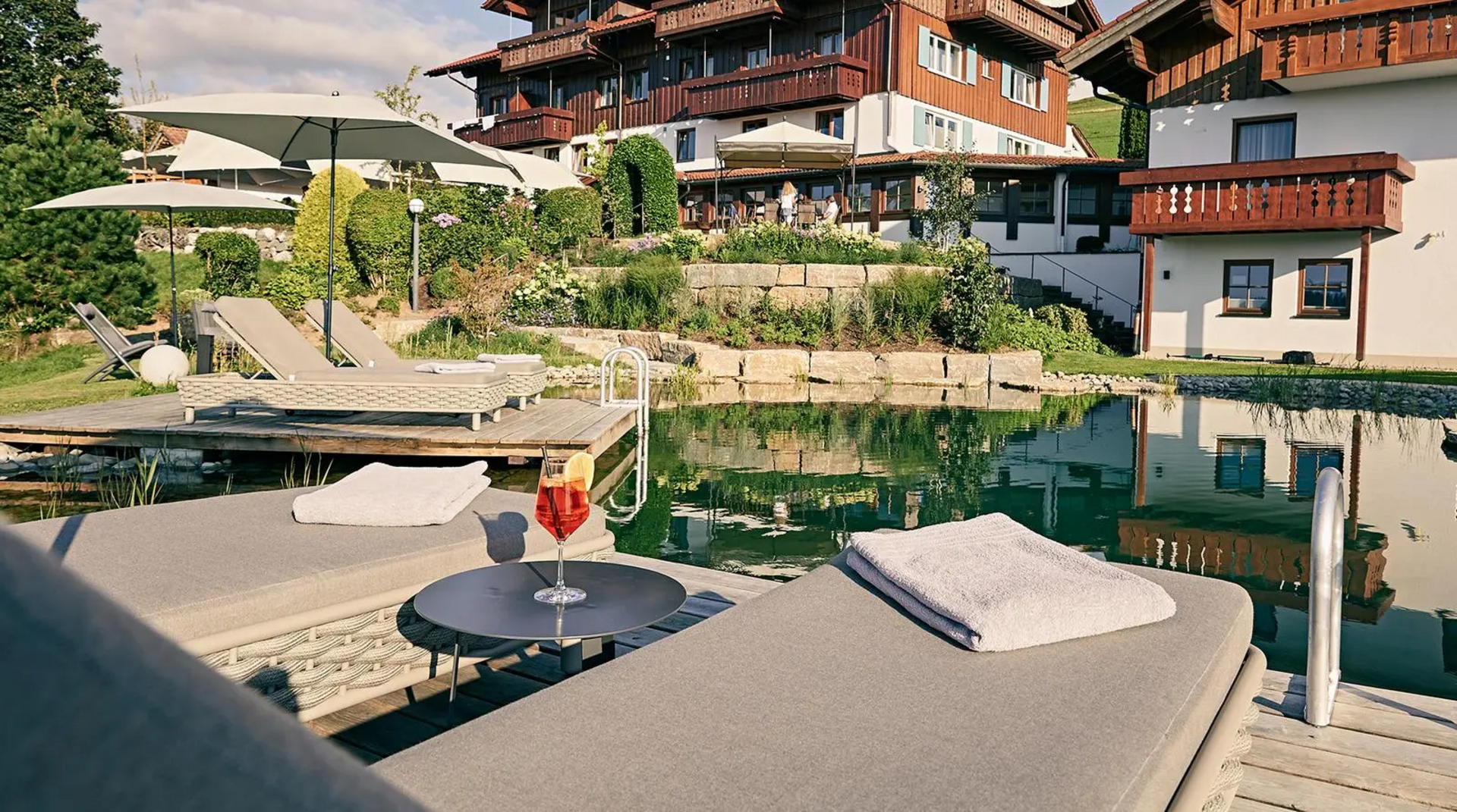 Liegen mit Handtüchern und einem Cocktail am Naturschwimmteich, im Hintergrund Hotel Birkenhof mit Sonnenschirmen und Gartenanlage.
