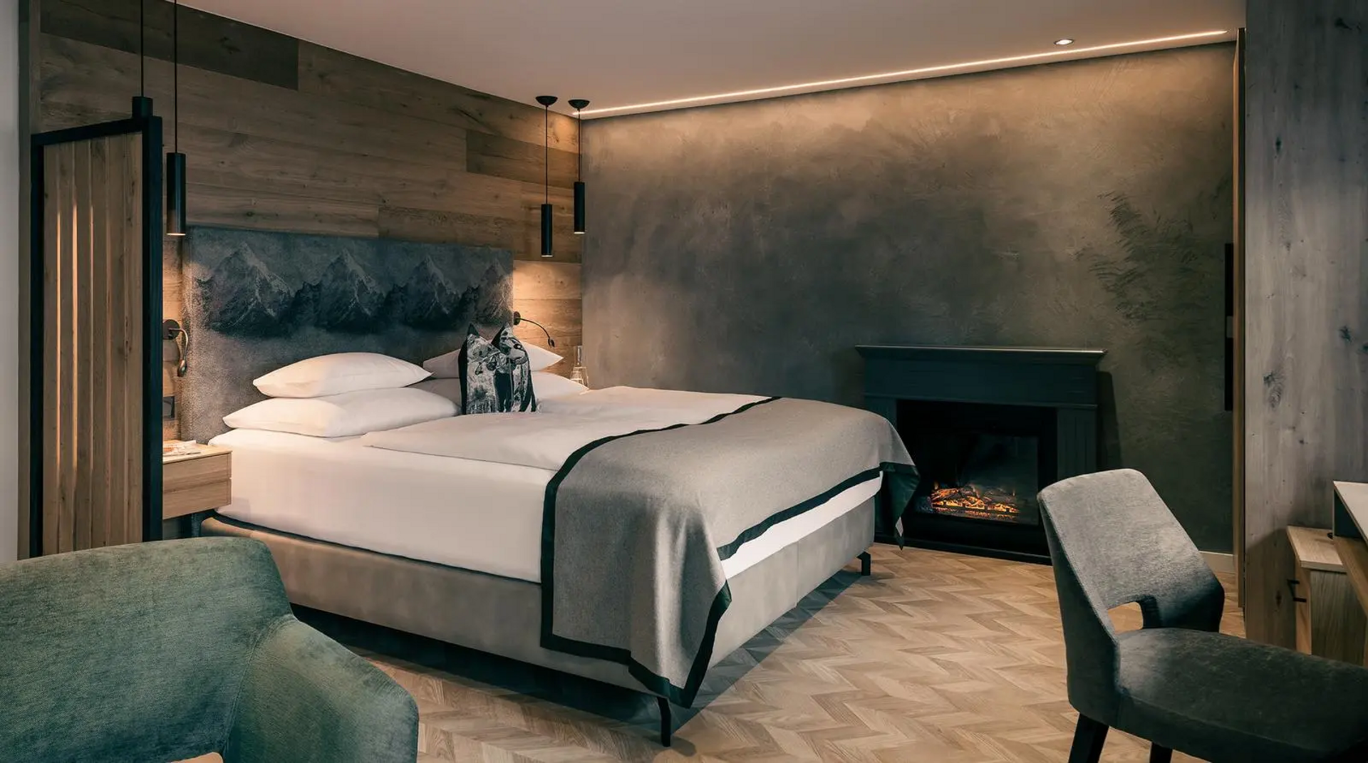 Modernes Doppelzimmer mit grauer Akzentwand, großem Bett, Kamin, Holz- und Stoffelementen sowie dezentem, indirektem Licht.
