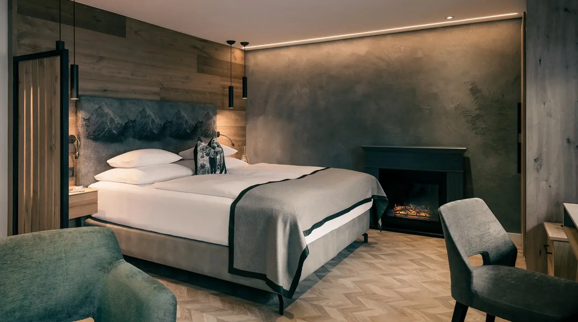 Modernes Doppelzimmer mit grauer Akzentwand, großem Bett, Kamin, Holz- und Stoffelementen sowie dezentem, indirektem Licht.
