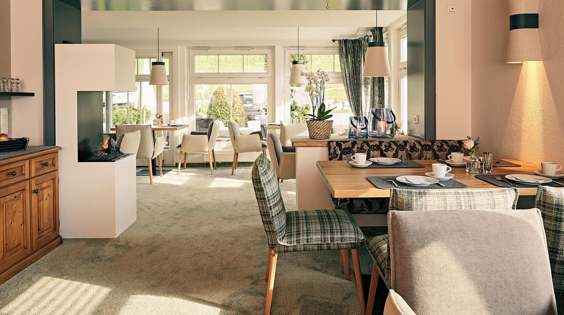 Heller Frühstücksraum im Restaurant des Hotel Birkenhof Oberstaufen mit modernem Kamin, gedeckten Tischen und großen Fenstern mit Blick ins Grüne.
