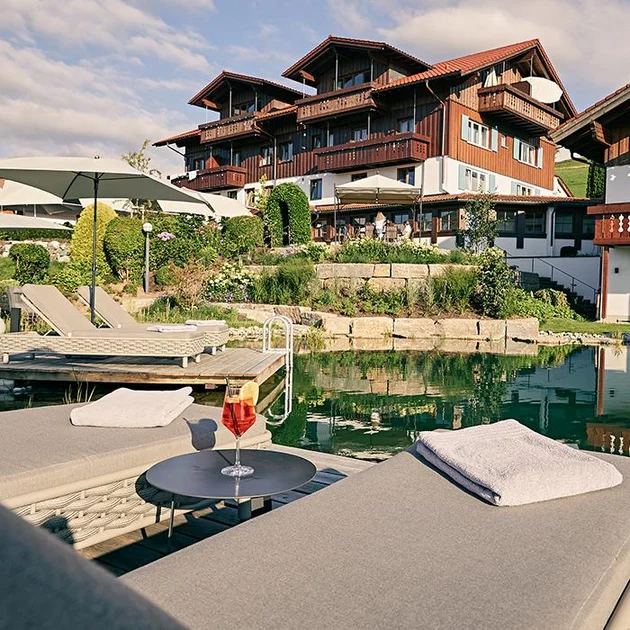 Blick vom Naturschwimmteich und Ruheliegen mit Aperol im Sommer auf das Hotel