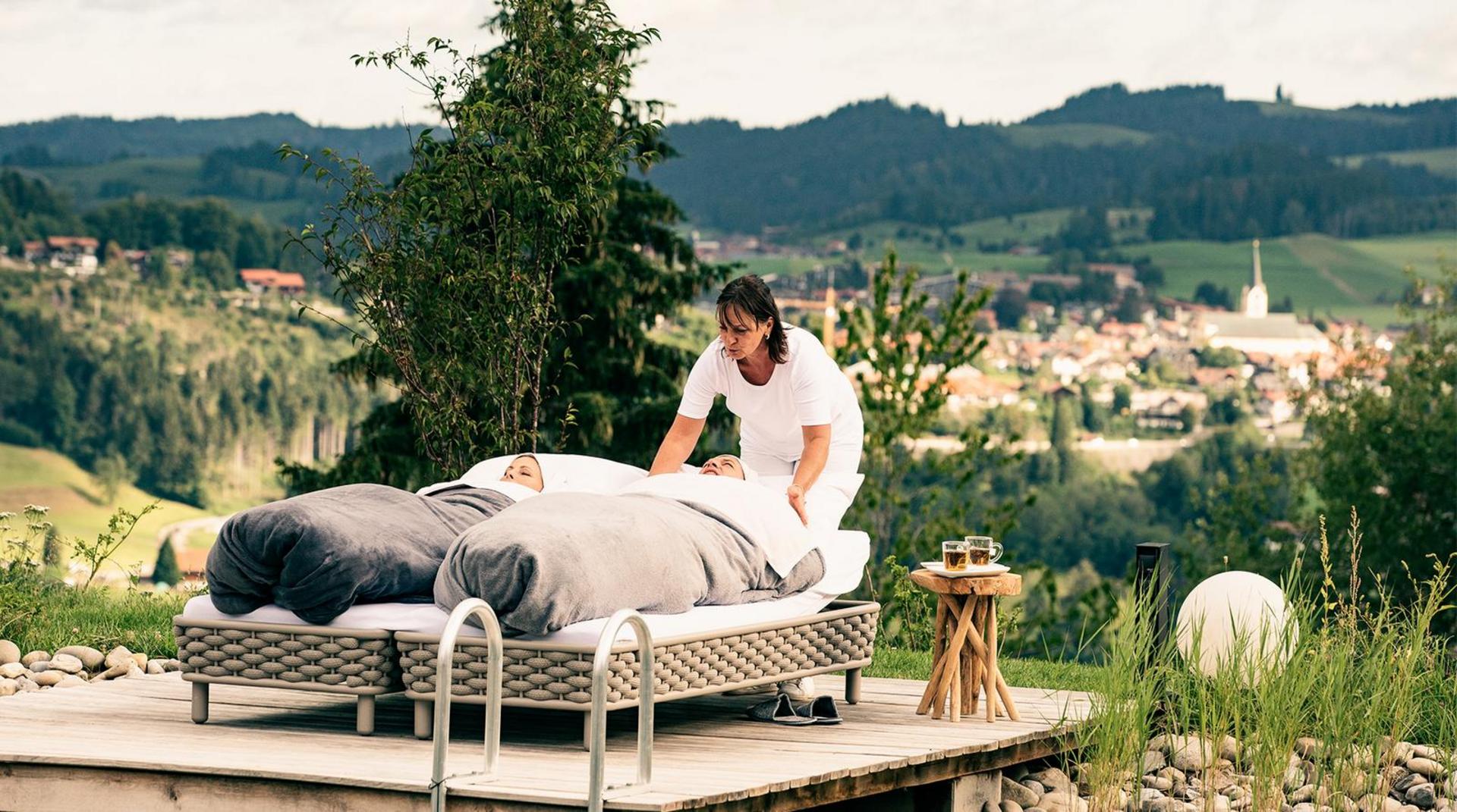 Frau wickelt Gast im Freien auf Liege in warme Decken für Schrothkur-Behandlung mit Blick auf die Allgäuer Hügellandschaft bei Oberstaufen
