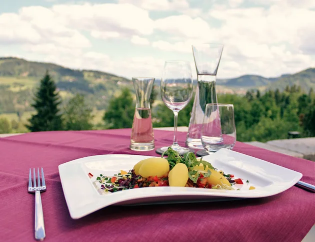 Gedeckter Tisch mit elegant angerichtetem Gericht und Gläsern, im Hintergrund Ausblick auf grüne Hügel und Berge.