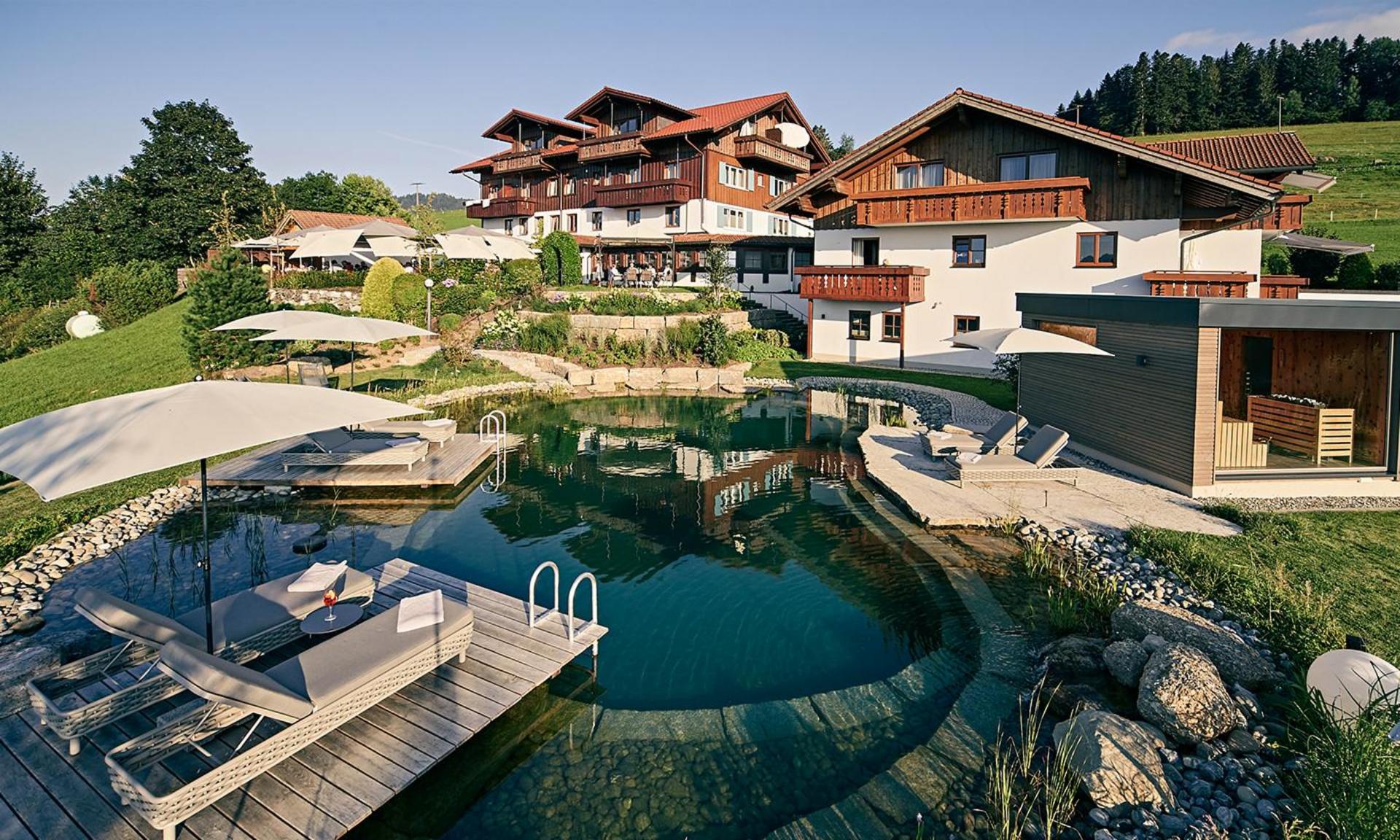 Weitläufige Hotelanlage des Hotel Birkenhof in Oberstaufen mit Naturschwimmteich, Holzdeck und gemütlichen Liegeflächen inmitten grüner Natur