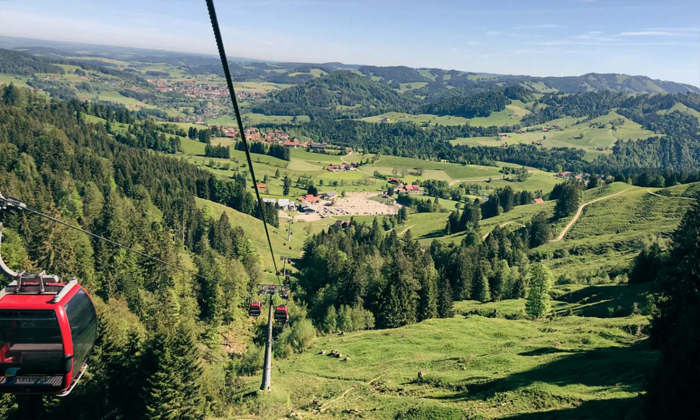 Rote Gondelbahn fährt über grüne Hügel und Wälder hinab ins Tal mit verstreuten Häusern und weitem Blick über die Allgäuer Landschaft.