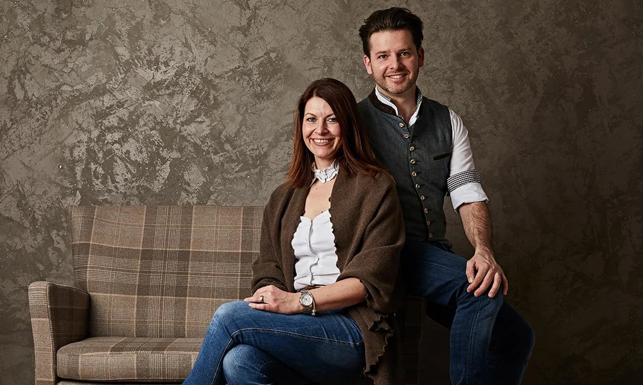 Porträt von Andrea und Thomas, den Gastgebern des Hotel Birkenhof in Oberstaufen, sitzend vor einer gemusterten Wand auf einem karierten Sofa