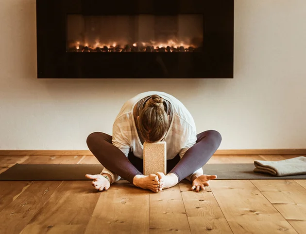 Frau übt Yoga im Sitzen vor einem Kaminfeuer, Stirn auf einem Yogablock abgestützt, Hände entspannt nach außen geöffnet.