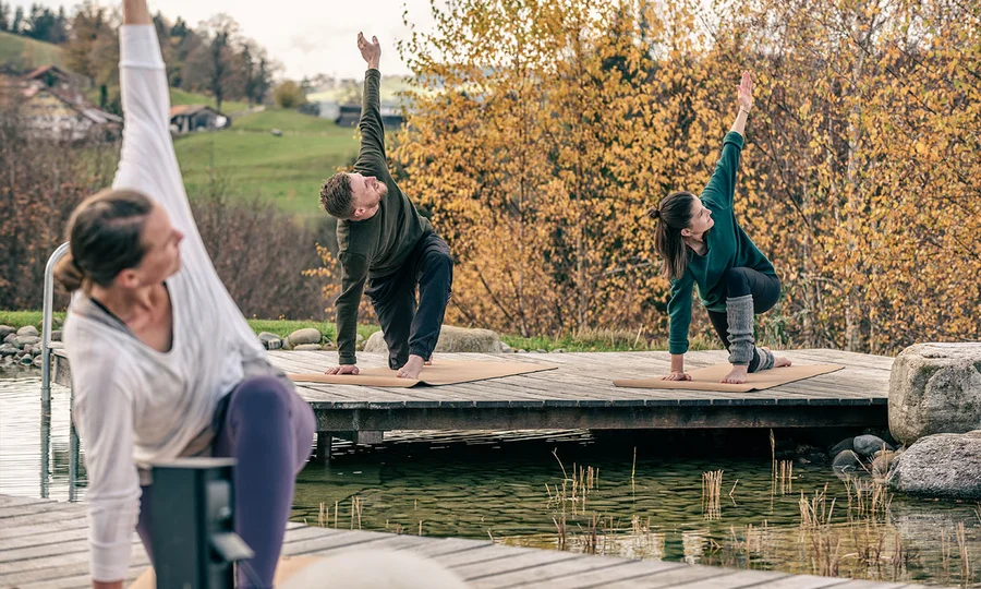 Drei Personen üben Yoga-Positionen auf Holzstegen am Birkenhof-Schwimmteich im Herbst.