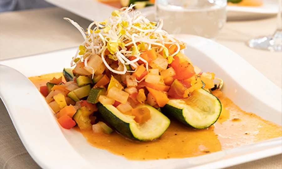 Zucchinihälften mit buntem Gemüse und Sprossen auf pikanter Sauce angerichtet.