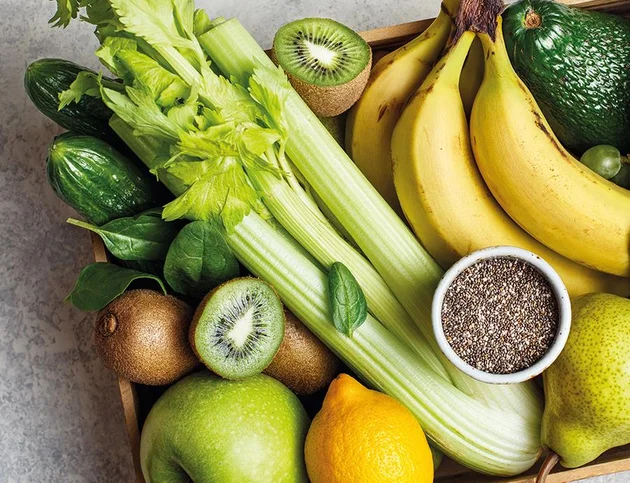 Holzkiste mit frischem Obst und Gemüse: Bananen, Kiwi, Zitrone, Apfel, Avocado, Sellerie, Gurken, Spinat und Schale mit Chiasamen.