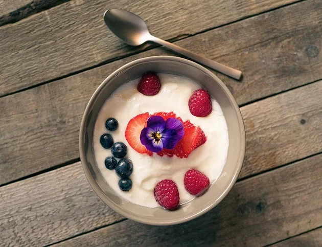 Schale Joghurt mit Beeren und essbarer Blüte, daneben ein Löffel.