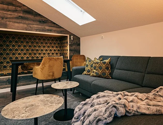 Gemütliche Sofaecke unter Dachschräge mit grauem Sofa, gemusterten Kissen, Decke, zwei runden Beistelltischen und Schreibtisch mit gelbem Stuhl vor Holzwand.