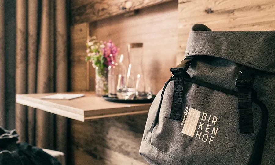 Moderner Hotelrucksack mit Birkenhof-Logo, daneben Holztisch mit Wassergläsern und Blumen.