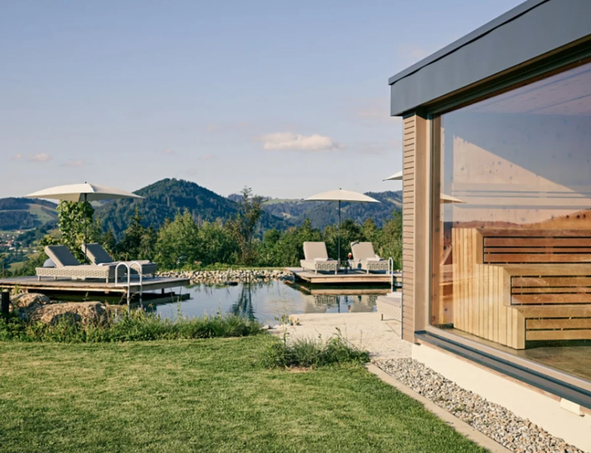 Wellnessbereich des Hotels Birkenhof in Oberstaufen mit Blick auf die Panoramasauna, Naturpool und Liegen mit Sonnenschirmen vor Bergkulisse.