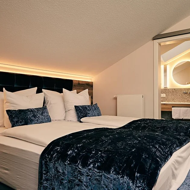 Gemütliches Schlafzimmer der Nagelfluhsuite im Hotel Birkenhof Oberstaufen mit großem Doppelbett, blauen Akzenten und Blick ins moderne Badezimmer.