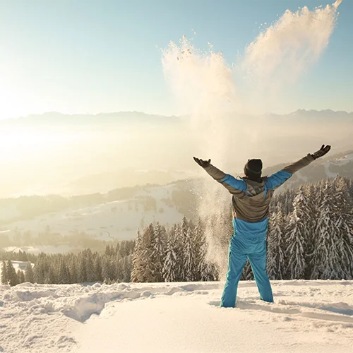 Mann im Skianzug wirft die Arme in die Höhe und steht freudig im verschneiten Allgäu mit Blick auf die Berge.
