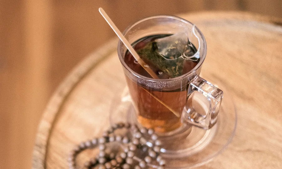 Glas mit heißem Kräutertee und Teebeutel auf Untertasse, daneben Holzperlen.