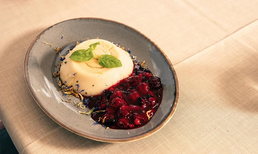 Dessert aus heller Creme mit Basilikumblatt garniert, serviert mit roter Beerensauce auf grauem Teller.