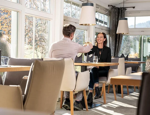 Paar stößt mit einem Glas Wein im hellen Restaurant des Hotel Birkenhof in Oberstaufen an