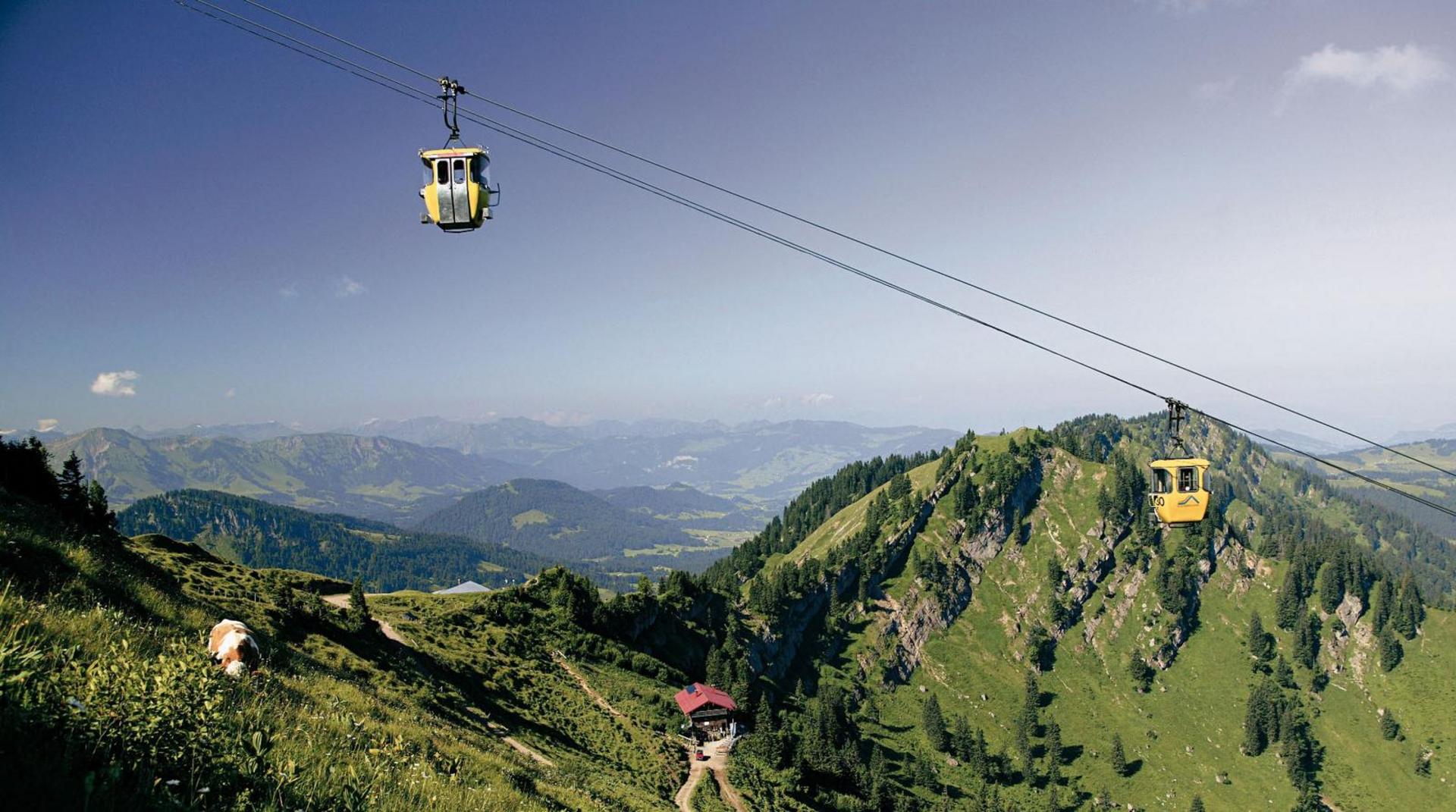 Gelbe Gondeln einer Seilbahn schweben über eine grüne Berglandschaft mit weitem Blick ins Allgäu.