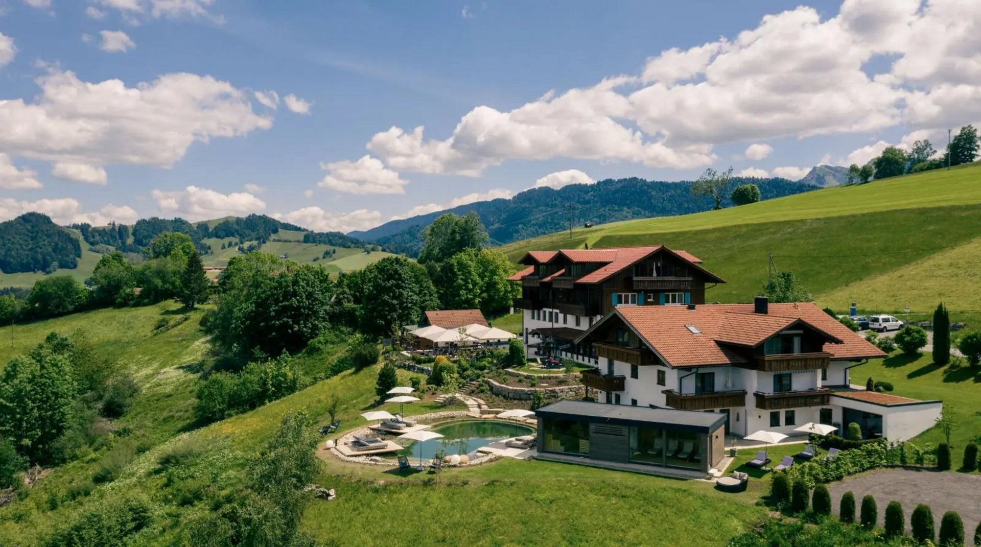 Hotel Birkenhof Oberstaufen in Alleinlage mit Panoramablick, umgeben von grünen Wiesen und Bergen, mit Natur-Schwimmteich im Vordergrund.