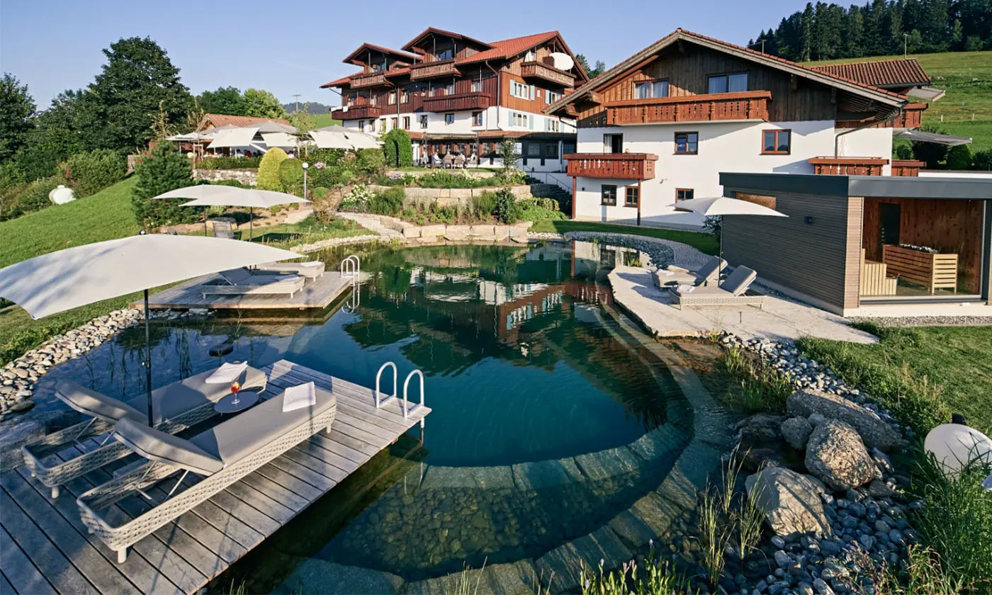 Großer Natur-Schwimmteich mit Holzliegedecks und Blick auf das Hotel Birkenhof in sonniger Hanglage