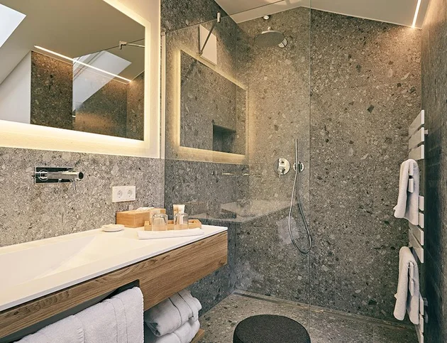 Modernes Badezimmer mit grauen Natursteinwänden, bodengleicher Dusche, beleuchtetem Spiegel, Holzwaschtisch und Handtuchheizkörper.