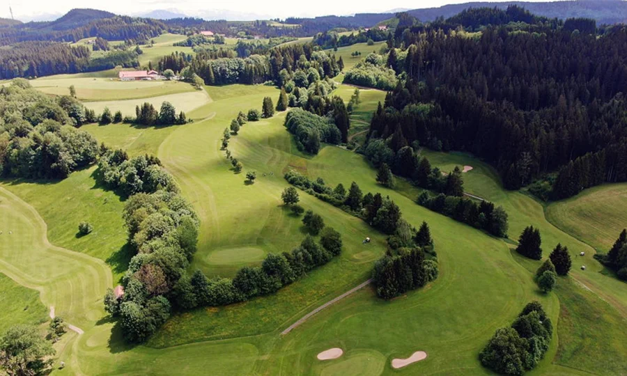 Luftaufnahme eines hügeligen Golfplatzes, umgeben von Wald und Wiesen.