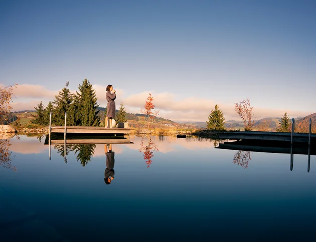 Person im Bademantel steht auf einem Steg am ruhigen See, im Hintergrund Berge und Bäume, Spiegelung im Wasser.