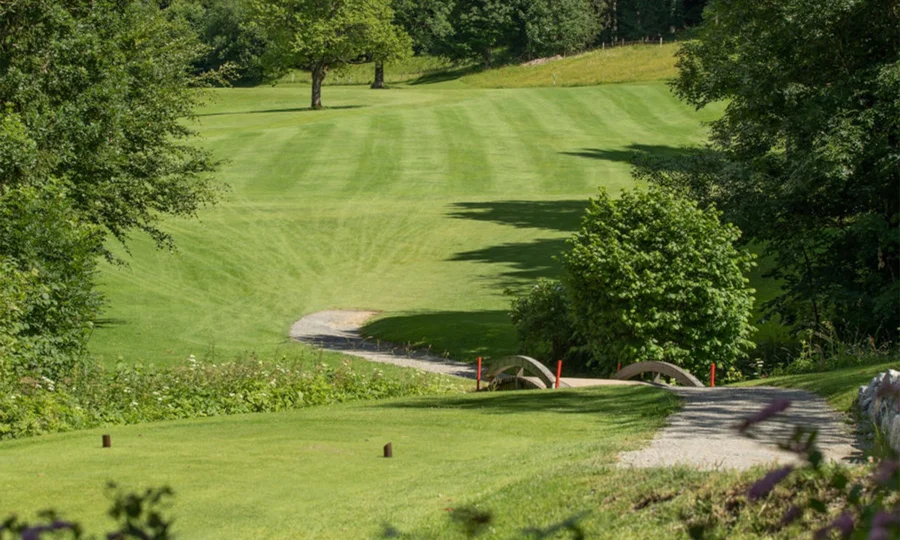 Schmaler Golfplatzabschnitt mit gepflegtem Rasen, Bäumen und kleinem Holzsteg.