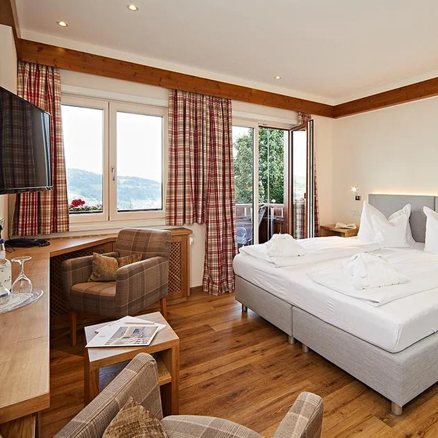 Helles Doppelzimmer im Hotel Birkenhof Oberstaufen mit Doppelbett, Sessel, Schreibtisch, Fernseher und Zugang zum Balkon mit Ausblick.