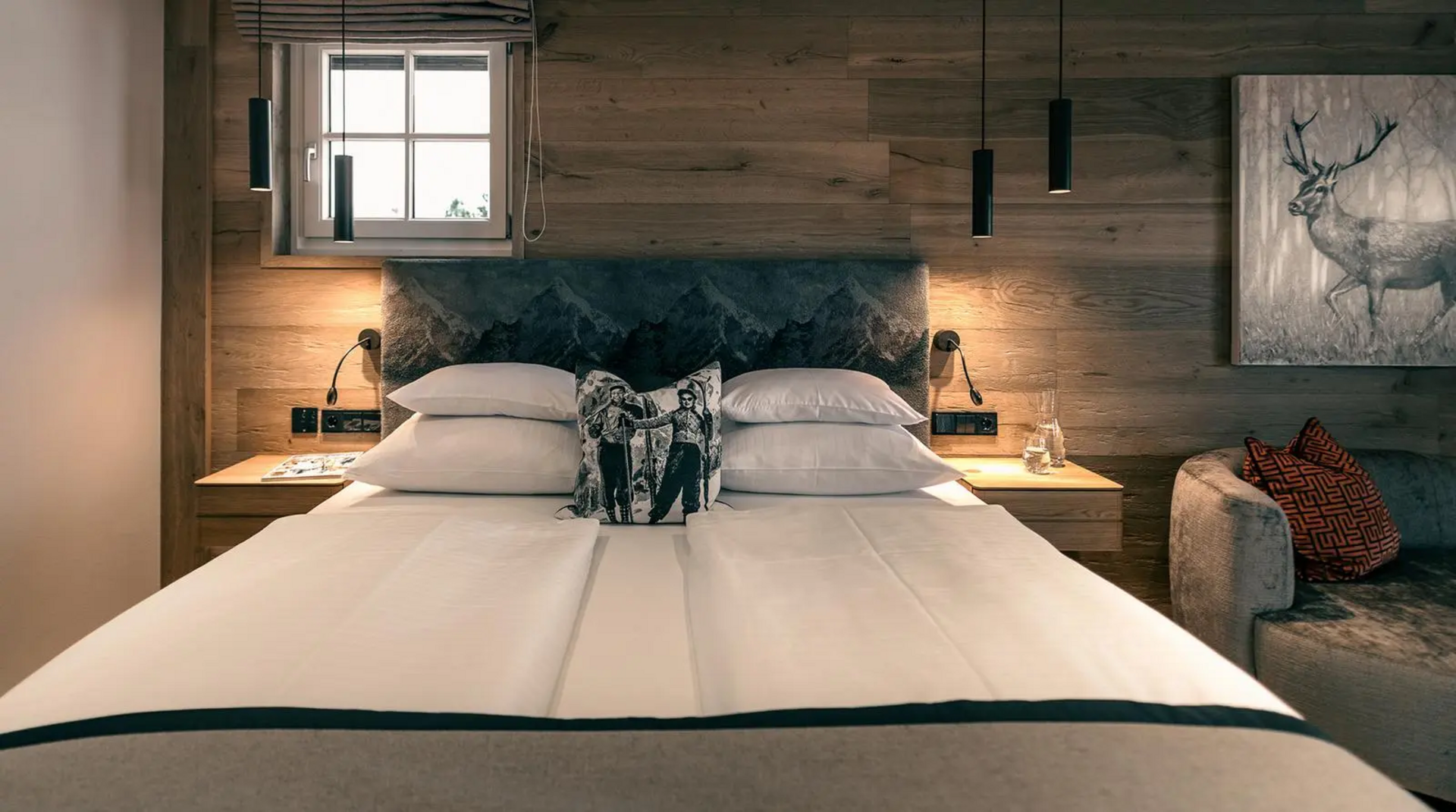 Doppelzimmer mit Holzverkleidung, großem Bett, Hirschmotiv-Kissen, seitlicher Couch und Wandbild eines Hirsches.