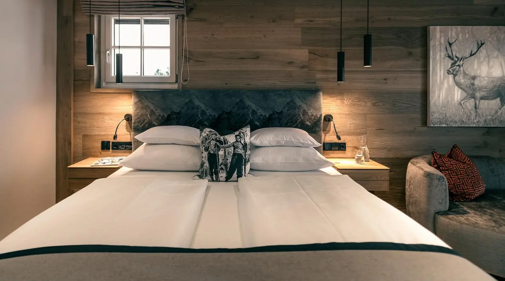 Doppelzimmer mit Holzverkleidung, großem Bett, Hirschmotiv-Kissen, seitlicher Couch und Wandbild eines Hirsches.