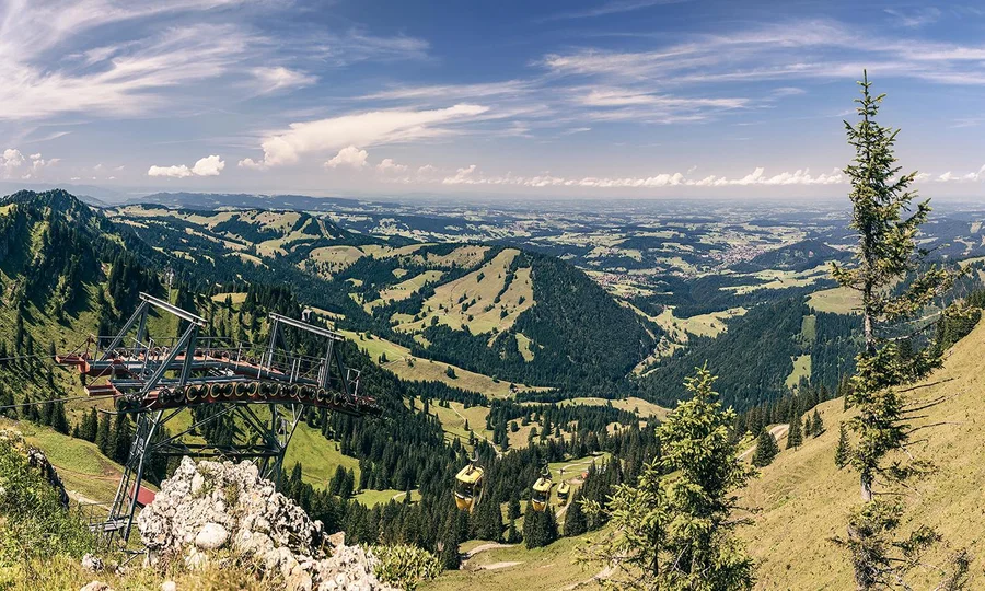 Panoramablick von den Bergen auf grüne Täler, Wälder und eine Bergbahnstation im Allgäu bei klarem Himmel.