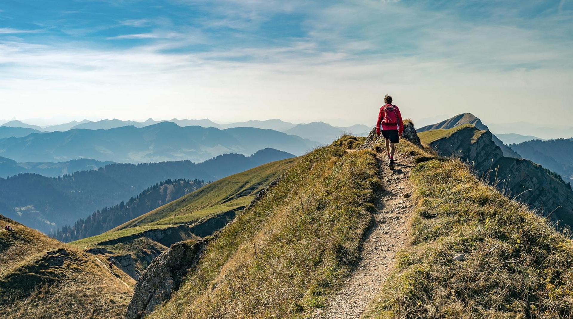 Wanderer mit rotem Rucksack geht auf einem schmalen Bergpfad, umgeben von grünen Gipfeln und weitem Blick ins Allgäu.