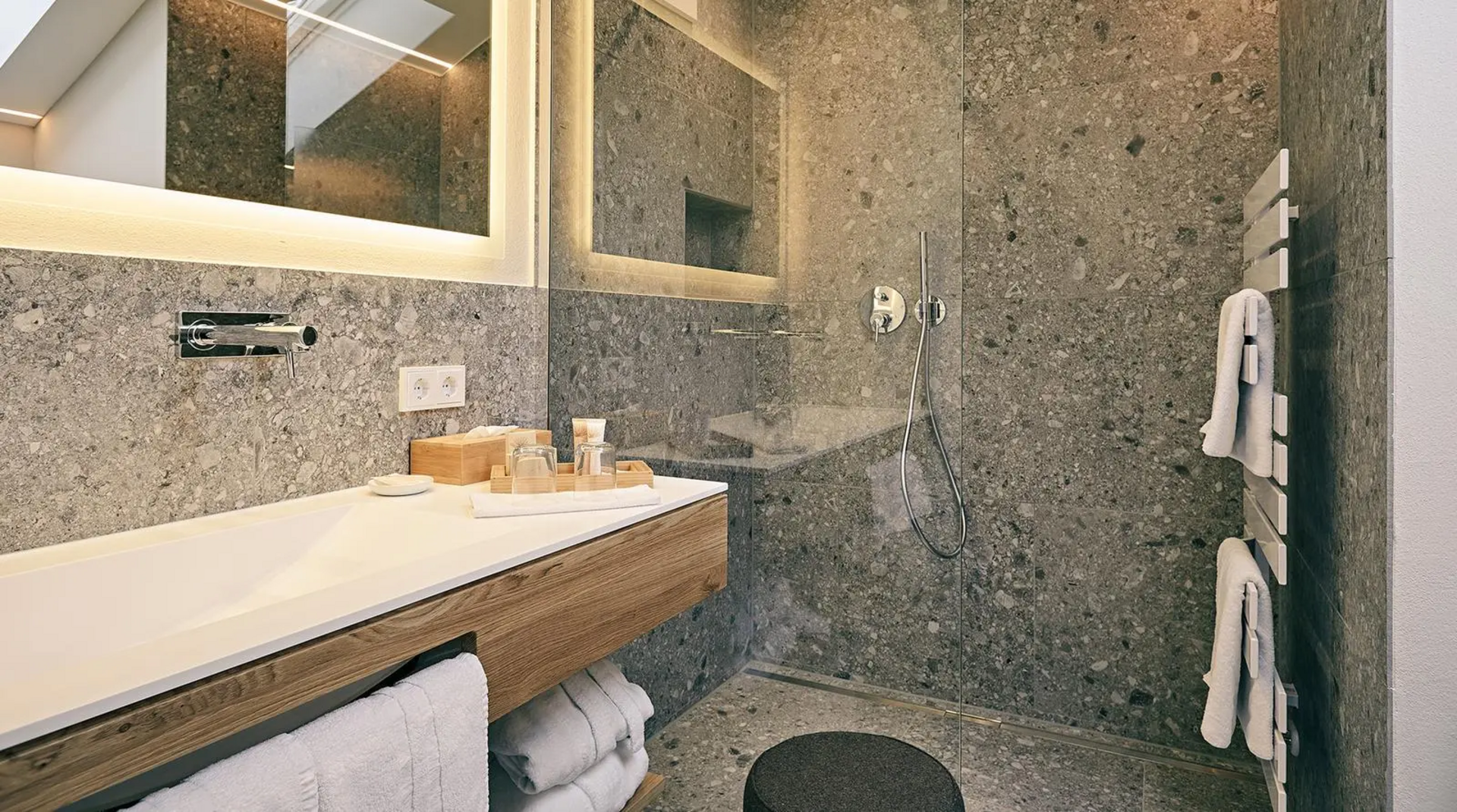 Modernes Badezimmer mit grauen Natursteinwänden, bodengleicher Dusche, beleuchtetem Spiegel, Waschtisch aus Holz und Handtuchheizkörper.