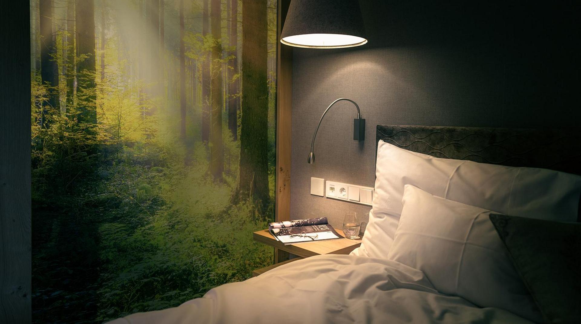 Bett mit Waldfotomotiv an der Wand und Nachttischleuchte.