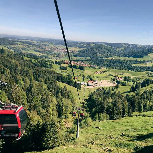 Fahrt mit der Hündlebahn in Oberstaufen mit roten Gondeln, die über grüne Wälder und Wiesen hinweg ins Tal mit weitem Panoramablick führen.