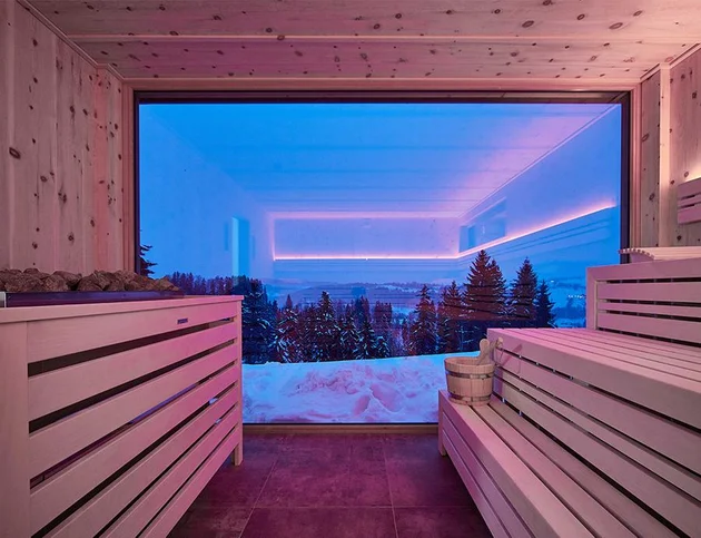 Moderne Sauna mit großen Panoramafenstern, die einen Blick auf verschneite Bäume und eine winterliche Landschaft bieten, innen mit hellem Holz verkleidet und stimmungsvoller LED-Beleuchtung in Rosa und Blau.