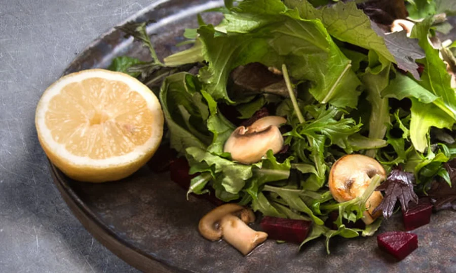 Grüner Blattsalat mit Champignons, Roter Bete und halber Zitrone auf dunklem Teller.