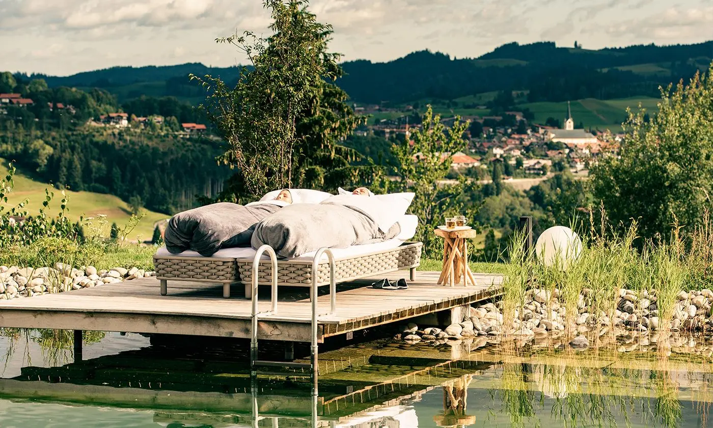 Zwei Personen liegen in Decken gehüllt auf Liegen am Steg eines Naturteichs, mit Blick auf die Landschaft und den Ort Oberstaufen.