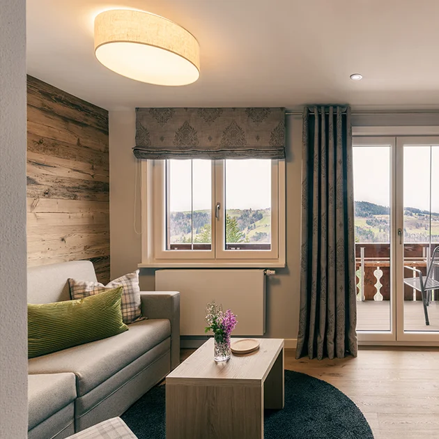 Helles Gartenapartment mit gemütlicher Sitzecke, Sofa und Couchtisch, Holzakzenten und großem Fenster mit Zugang zum Balkon und Ausblick auf die Berge.