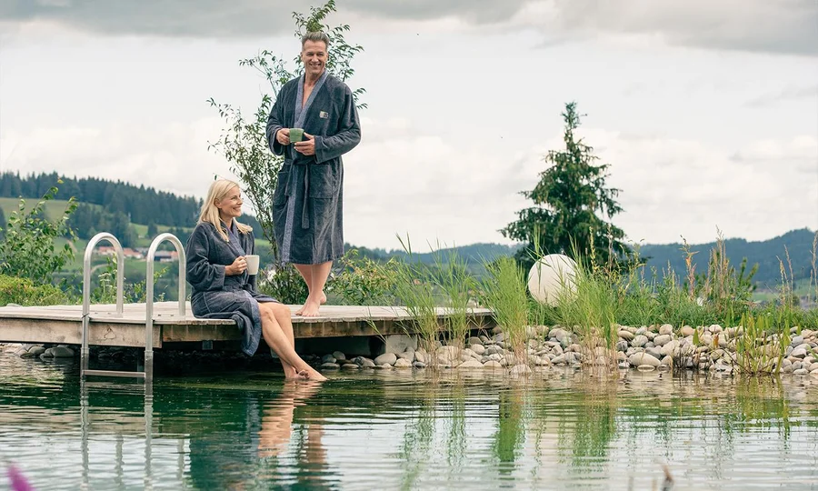 Mann und Frau im Bademantel sitzen und stehen auf einem Steg am Naturteich, beide mit Tassen in der Hand, umgeben von Natur und Bergen.