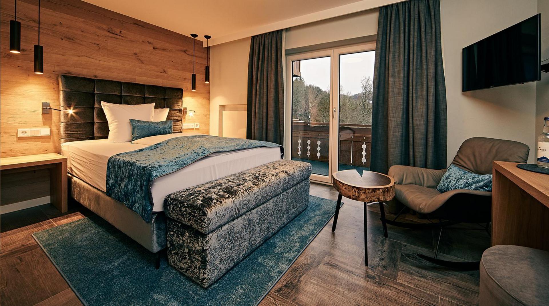 Stilvolles Einzelzimmer mit Boxspringbett, blau-grauen Textilien, Holz- und Polsterelementen, Sessel mit Beistelltisch sowie Zugang zum Balkon.