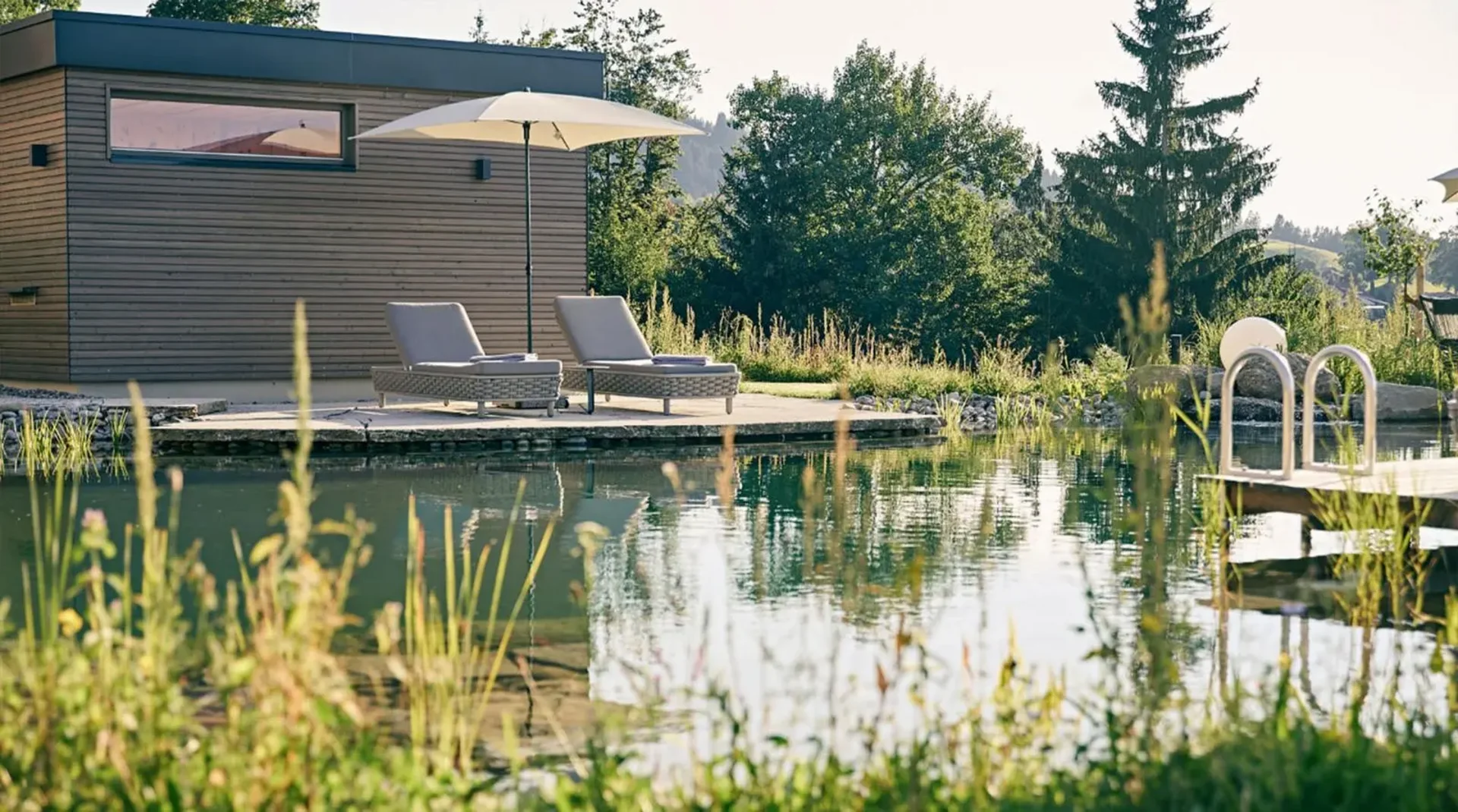 Zwei Liegen mit Sonnenschirm an einem Holzdeck direkt am Naturschwimmteich, umgeben von Grünpflanzen und Bäumen.
